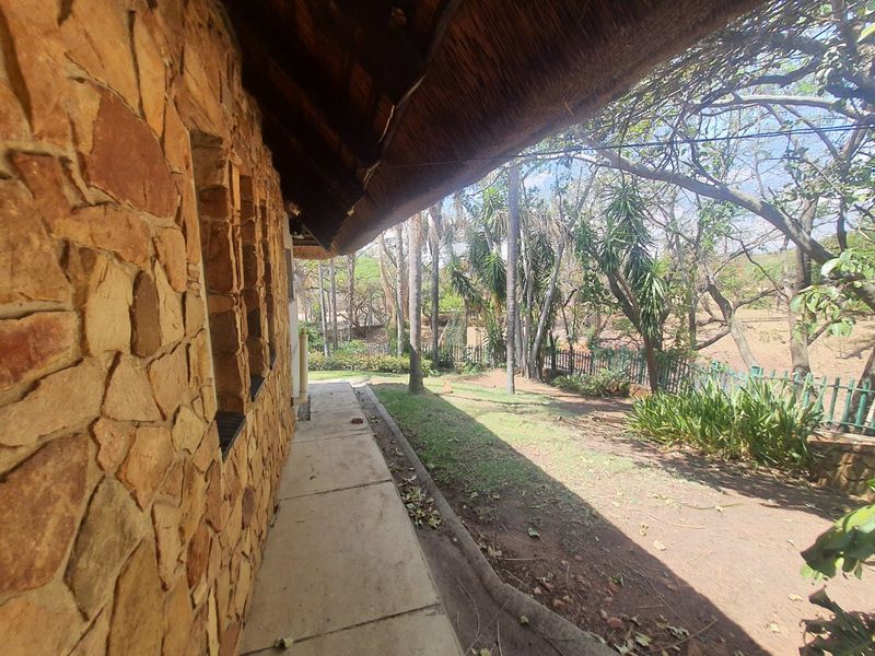 A covered patio area featuring rustic stone wall cladding and a traditional thatched roof, overlooking a garden with mature trees and perimeter fencing.