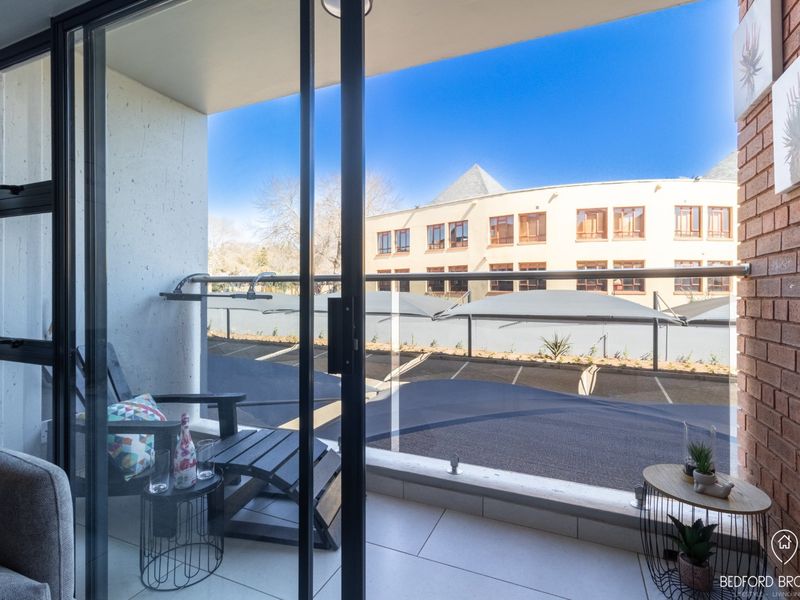 A modern balcony featuring a glass balustrade, tiled flooring, and an exposed brick wall, offering a view of the parking area.