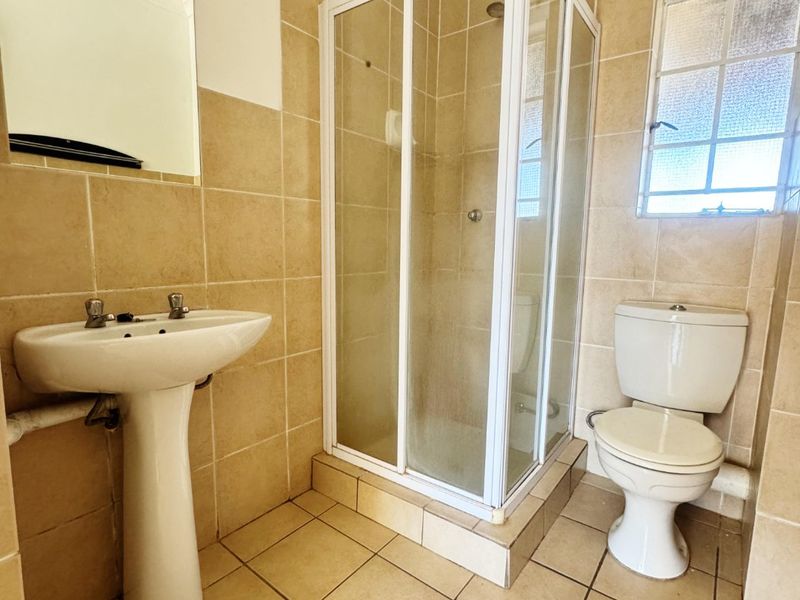 A functional bathroom featuring a pedestal basin, a corner shower enclosure, and a standard toilet with neutral wall and floor tiling.