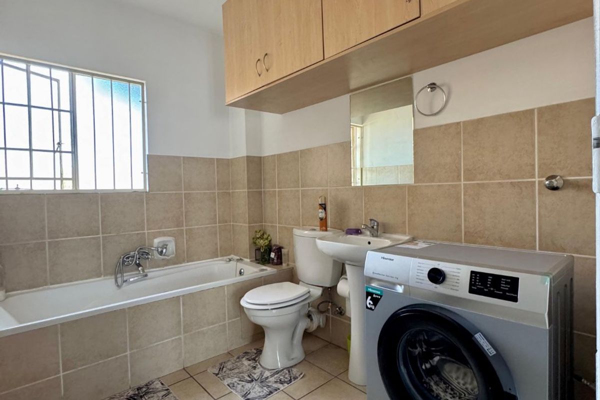 A functional bathroom featuring a bathtub, pedestal basin, toilet, and overhead cabinetry, with neutral tiled finishes and integrated laundry plumbing.