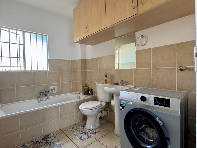 A functional bathroom featuring a bathtub, pedestal basin, toilet, and overhead cabinetry, with neutral tiled finishes and integrated laundry plumbing.