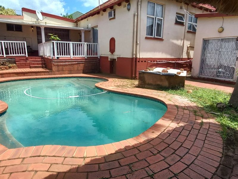 Rear garden area featuring a swimming pool, brick paved surround, and a raised patio with white balustrades attached to the main house structure.