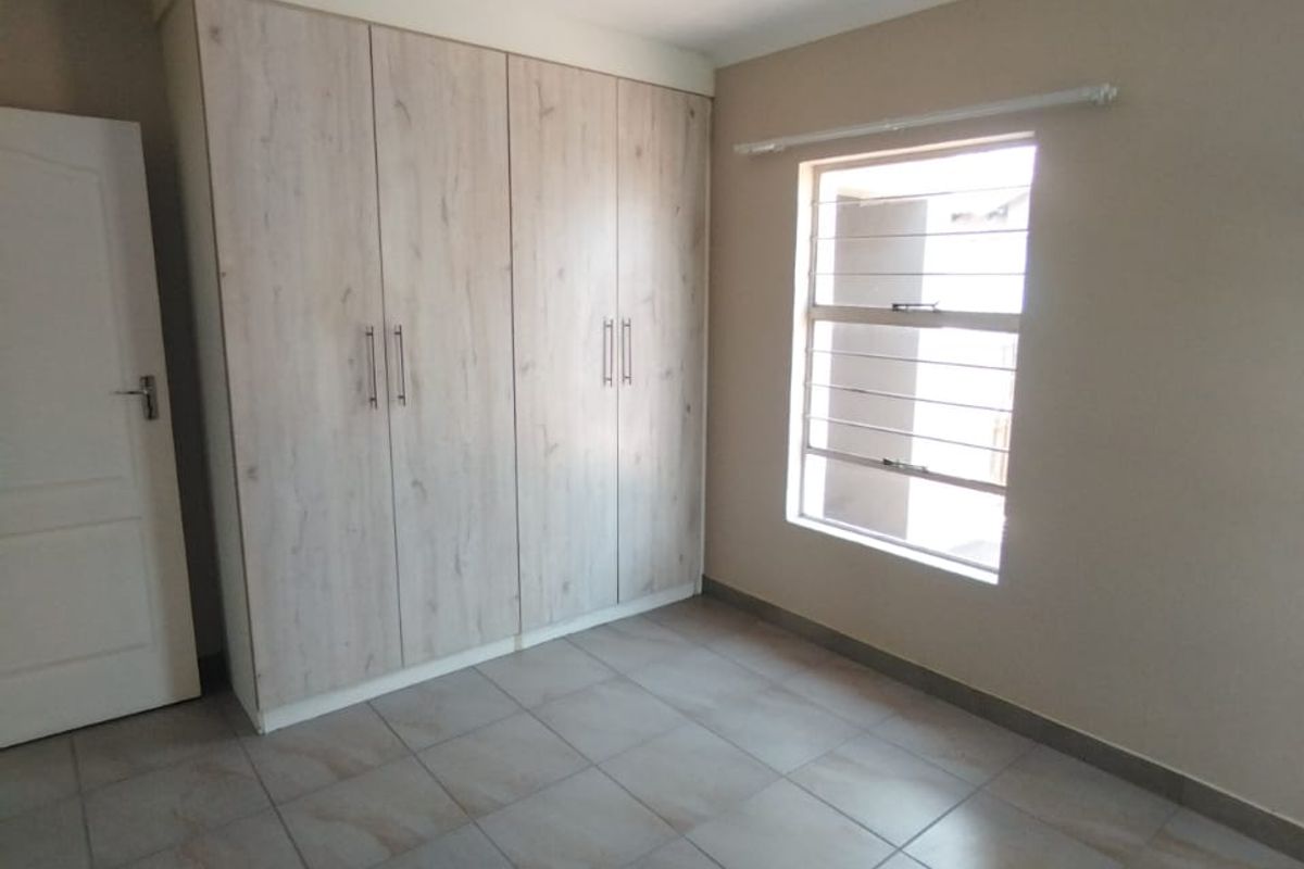 A clean, empty bedroom featuring neutral tiled flooring, built-in wooden cupboards, and a window fitted with security bars.