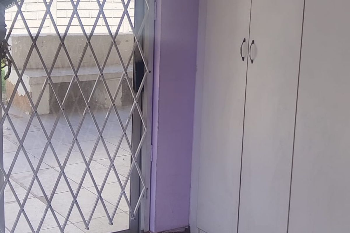 A bedroom interior featuring built-in cupboards, tiled flooring, and a sliding glass door with a security gate leading to an outdoor area.