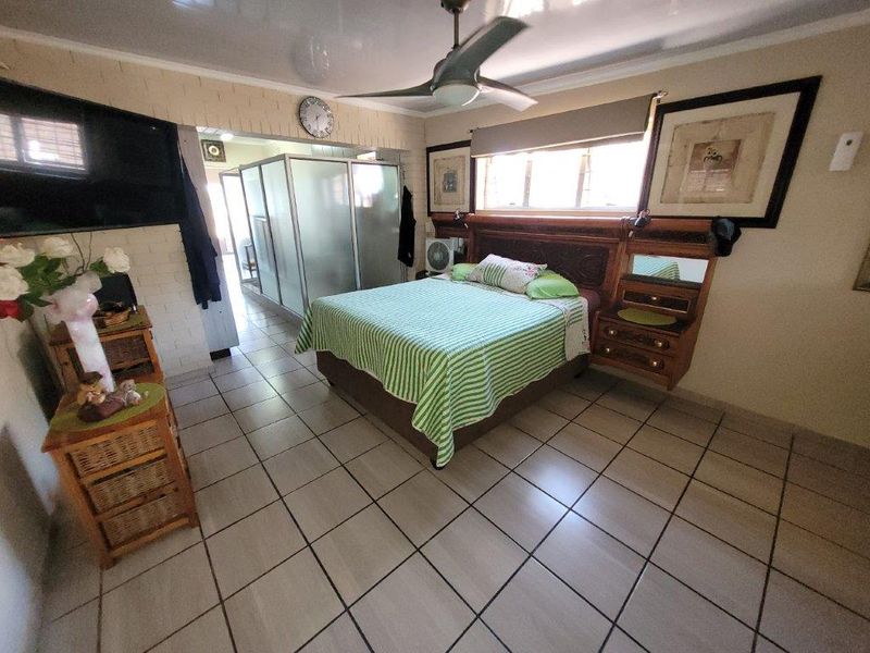 Spacious bedroom featuring tiled flooring, a ceiling fan, and a built-in headboard, with an adjoining en-suite bathroom area visible through a sliding door.