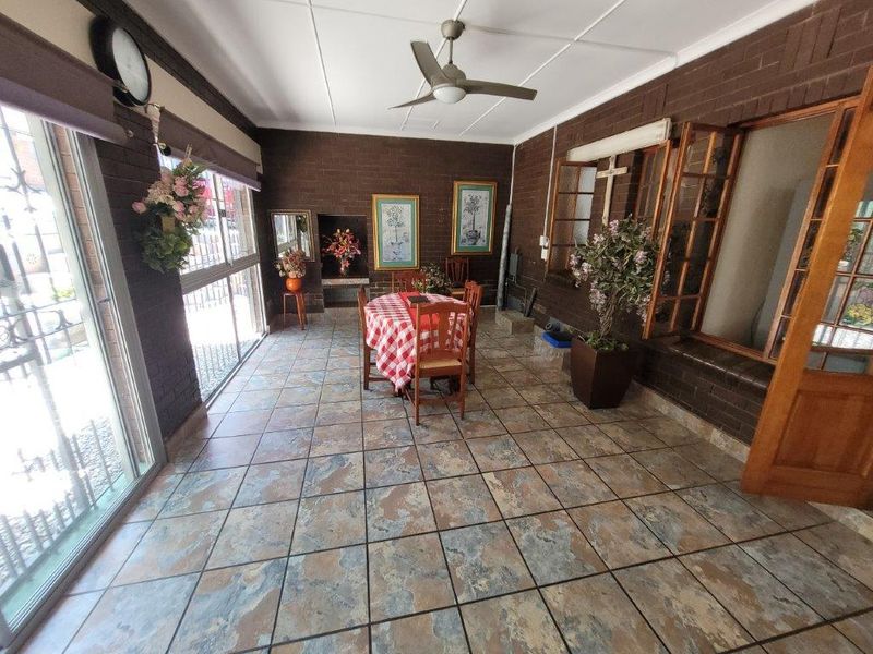A spacious enclosed patio featuring exposed brick walls, tiled flooring, a ceiling fan, and a built-in fireplace, offering a versatile indoor-outdoor living space.