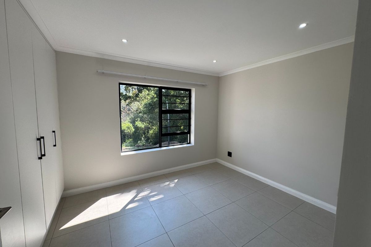 A bright, empty bedroom featuring neutral tiled flooring, built-in cupboards, recessed downlights, and a large window with a garden view.