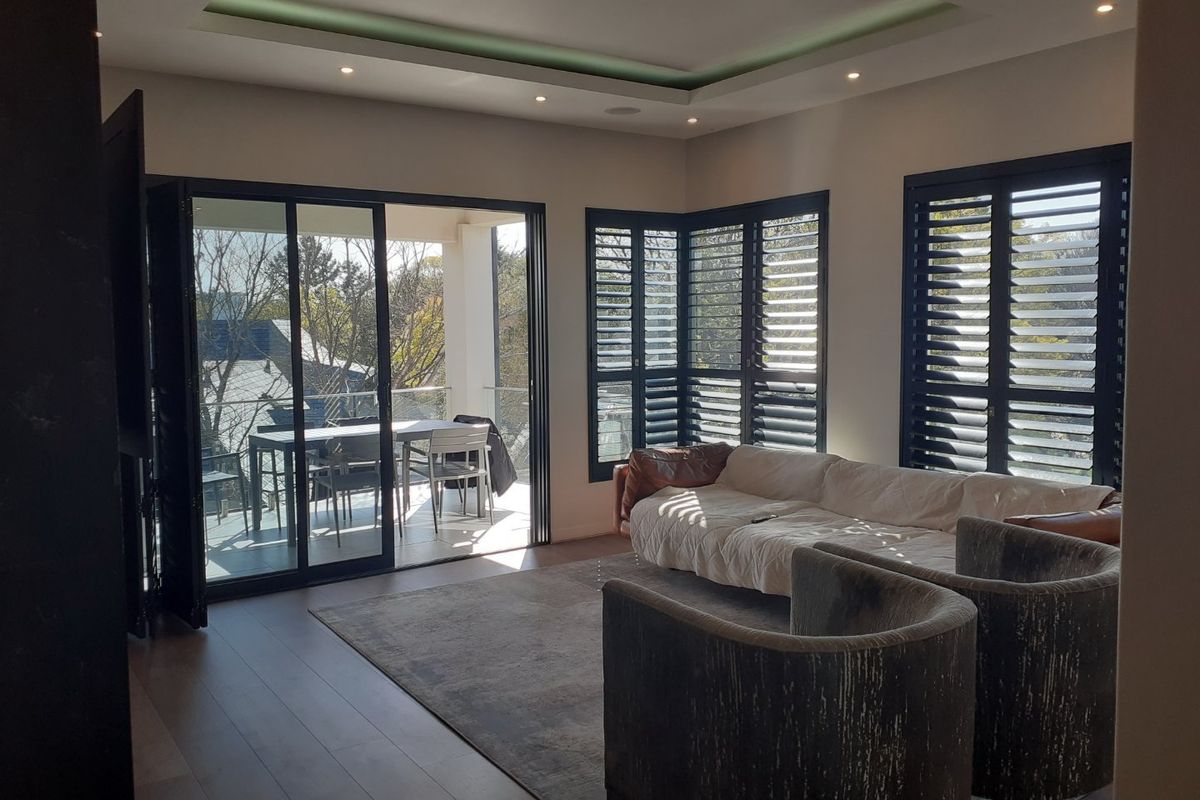 Spacious living room featuring timber flooring, recessed tray ceiling lighting, and large aluminium stacker doors opening onto a private balcony with plantation shutters.