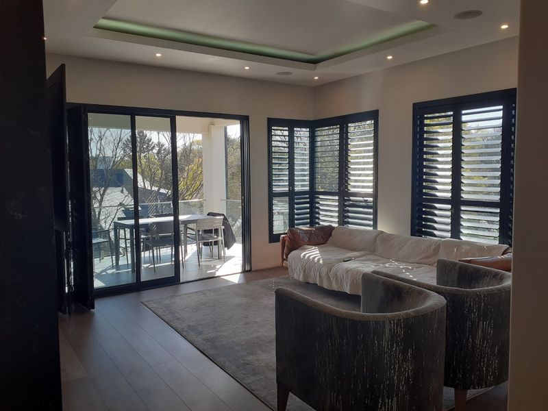 Spacious living room featuring timber flooring, recessed tray ceiling lighting, and large aluminium stacker doors opening onto a private balcony with plantation shutters.