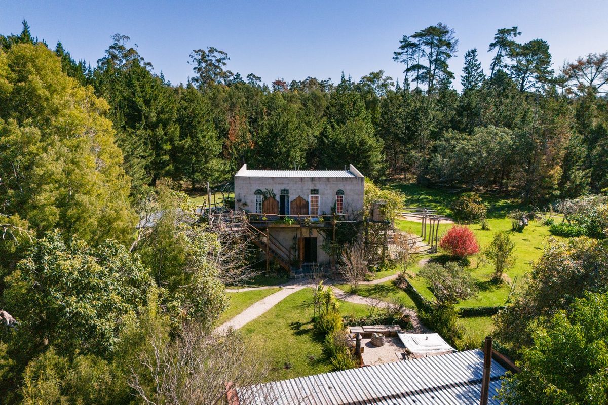 Aerial view of a rustic stone farmhouse set within a lush, wooded landscape, featuring a balcony, external staircase, and expansive garden grounds.