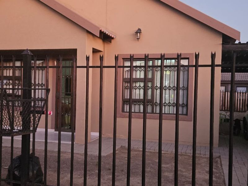 Front elevation of a single-story house featuring a paved driveway, metal security fencing, and security bars on windows.