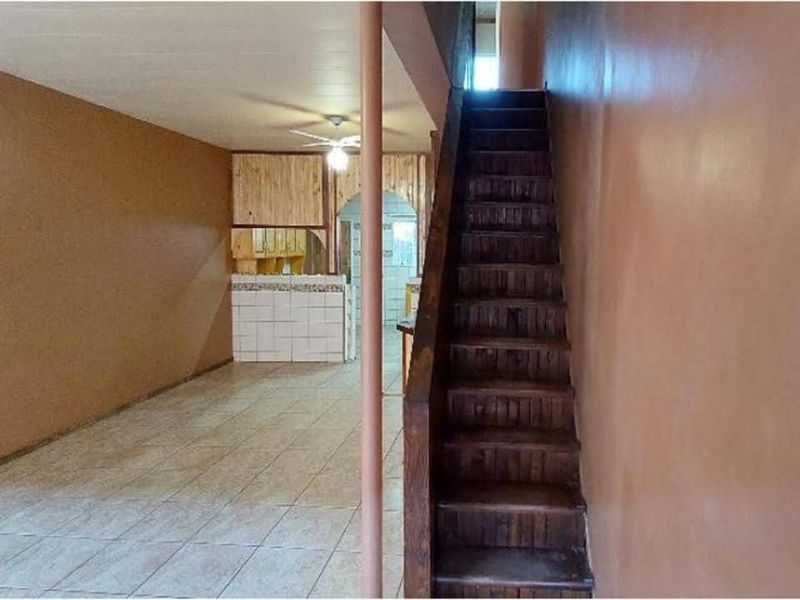 Open plan living area featuring tiled floors, a wooden staircase, and a dated kitchen area with built-in cabinetry.