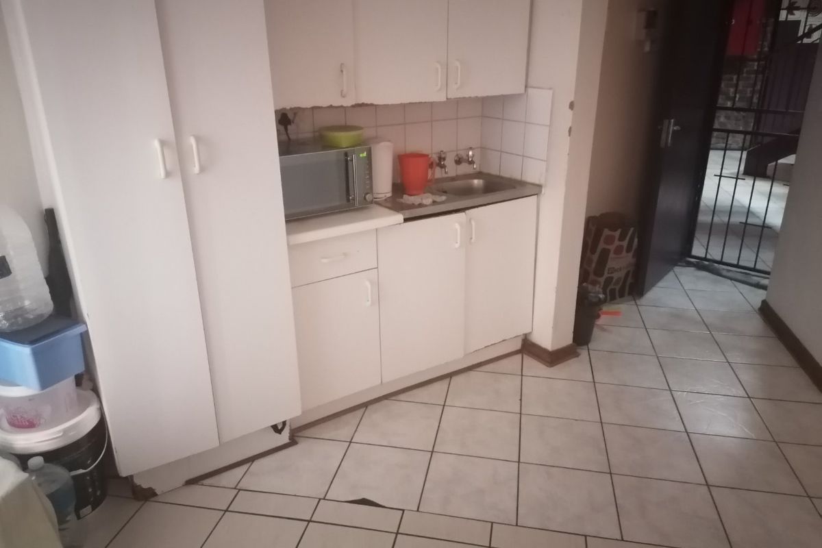 A compact kitchen area featuring white built-in cabinetry, a stainless steel sink, and tiled flooring, currently showing signs of wear and damage.