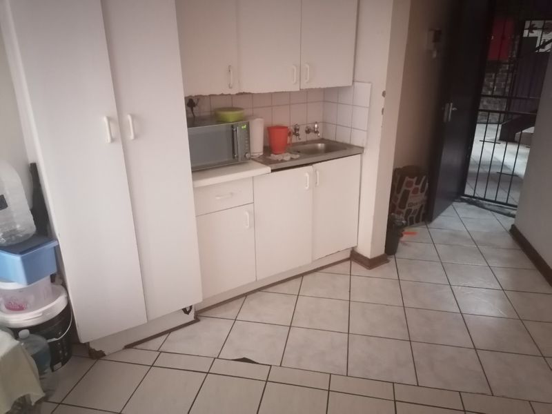 A compact kitchen area featuring white built-in cabinetry, a stainless steel sink, and tiled flooring, currently showing signs of wear and damage.