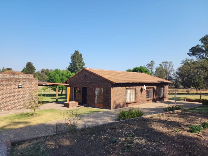 A rear view of a face brick farm house featuring a tiled roof, paved patio area, and established garden surroundings.