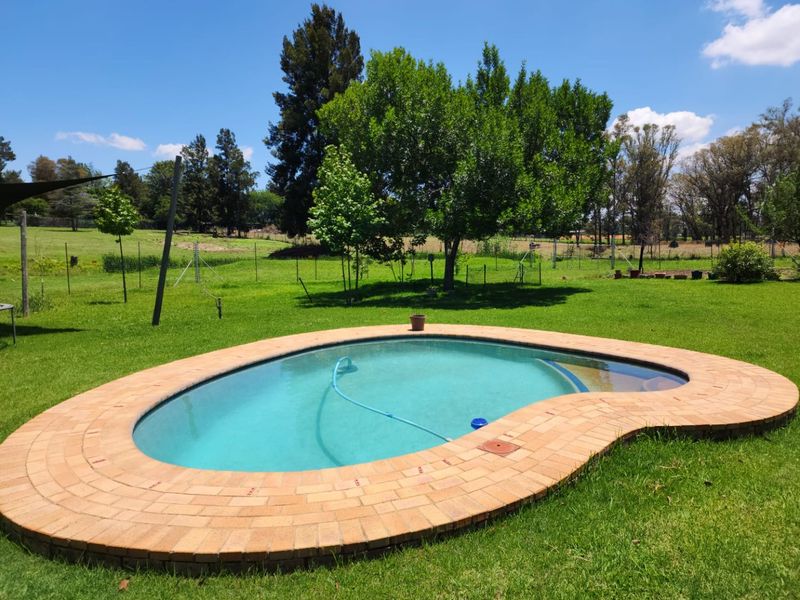 A kidney-shaped swimming pool with brick coping set within a spacious, well-maintained garden on a farm property.