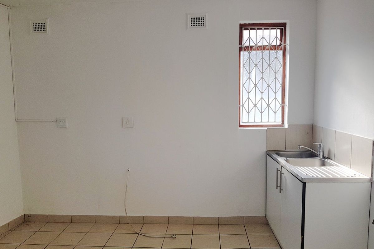 A compact, empty kitchen area featuring tiled flooring, a stainless steel sink unit, and a window with security bars.