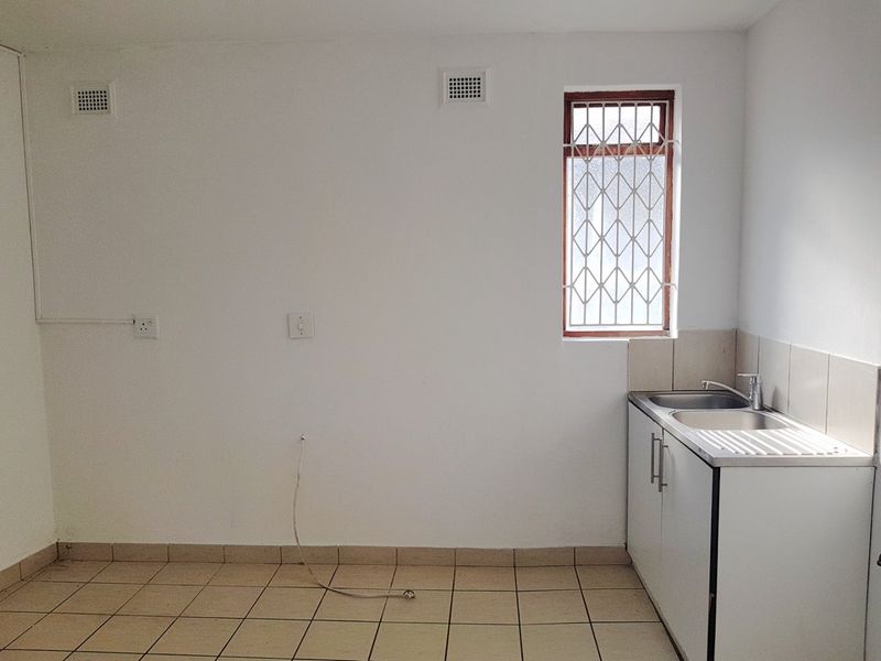 A compact, empty kitchen area featuring tiled flooring, a stainless steel sink unit, and a window with security bars.