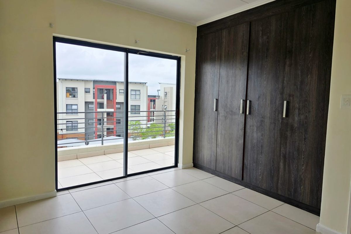 A bright, empty bedroom featuring modern dark wood built-in cupboards, large tiled flooring, and a sliding glass door leading to a private balcony.