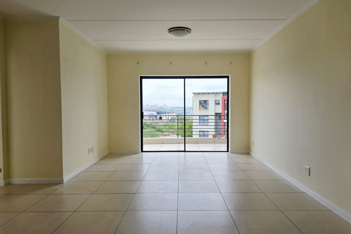 A bright, empty living room featuring neutral tiled flooring and a large sliding glass door that opens onto a balcony with urban views.