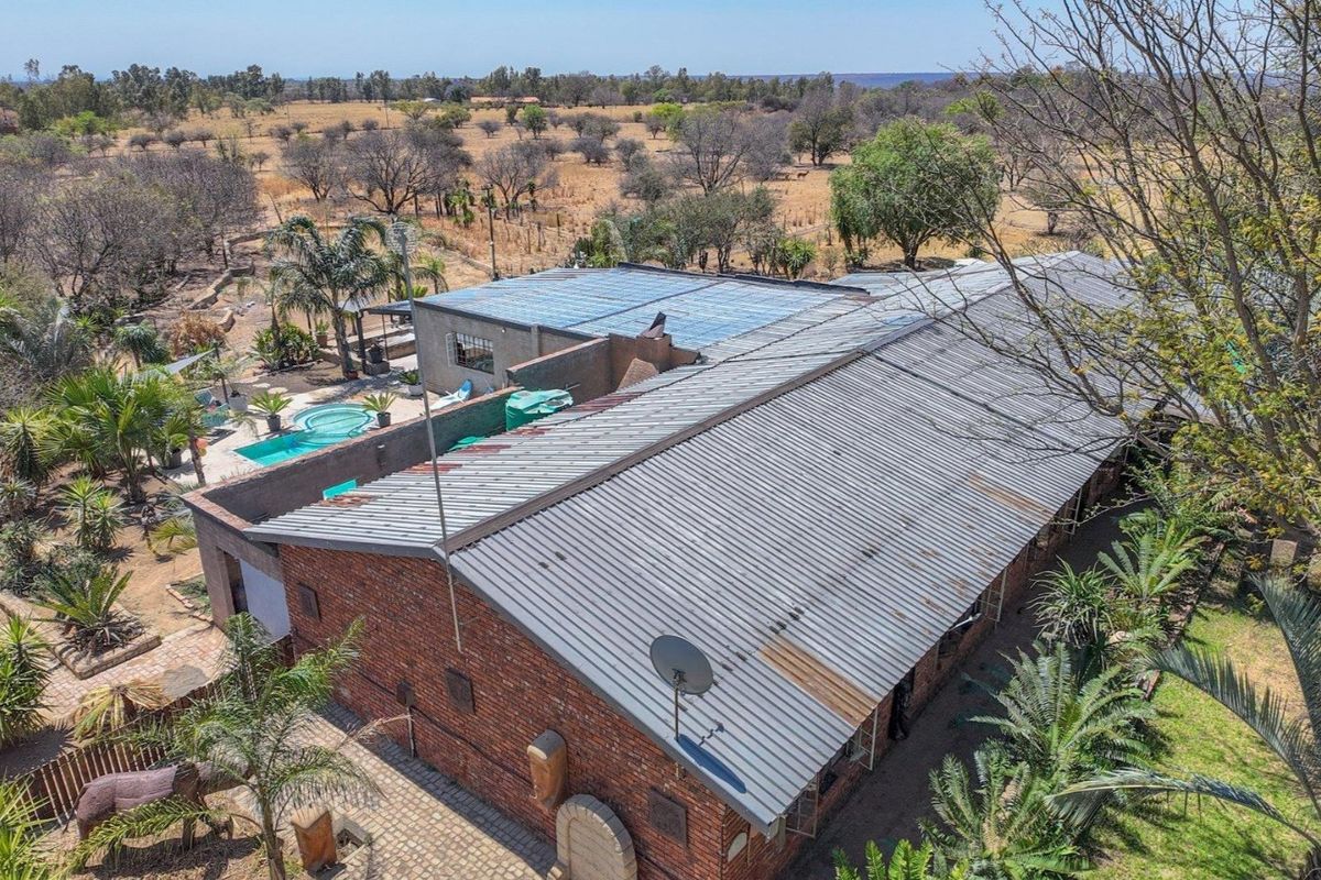 Aerial view of a farm property featuring a brick dwelling with a corrugated metal roof, an adjacent swimming pool, and surrounding rural landscape.