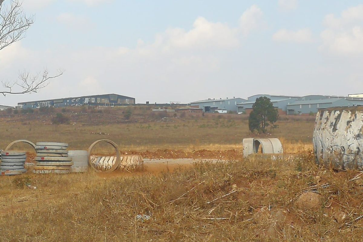 A view of an undeveloped residential plot featuring dry grass and scattered concrete drainage pipes with industrial buildings in the background.