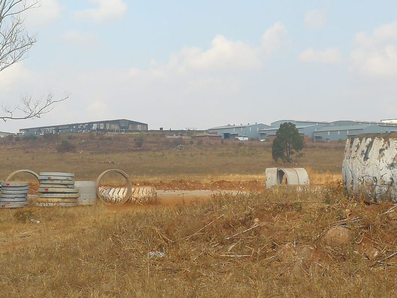 A view of an undeveloped residential plot featuring dry grass and scattered concrete drainage pipes with industrial buildings in the background.