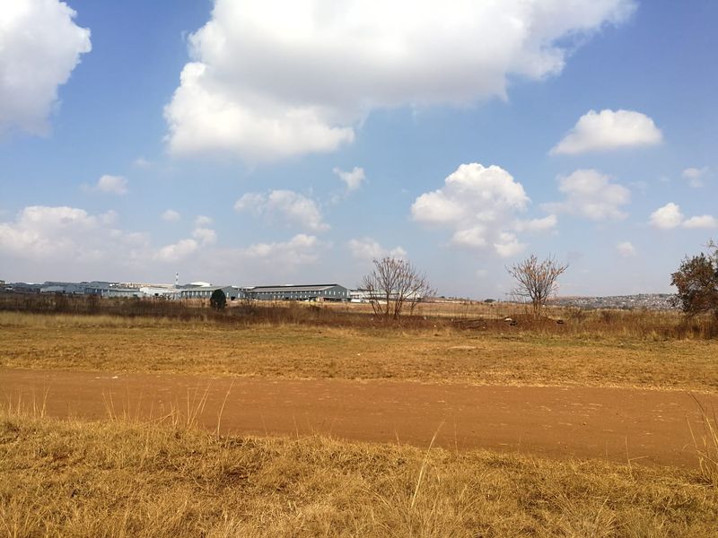 A wide-angle view of an undeveloped residential plot featuring dry grass and a dirt access road with distant industrial buildings in the background.