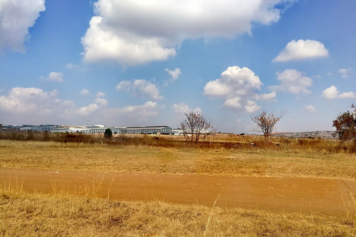 A wide-angle view of an open, undeveloped residential land plot with dry grass and distant industrial buildings under a clear blue sky.