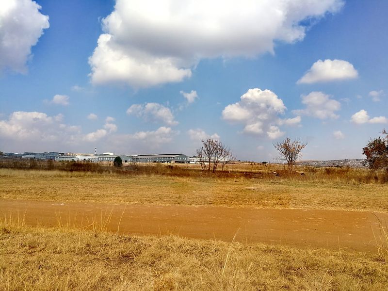 A wide-angle view of an open, undeveloped residential land plot with dry grass and distant industrial buildings under a clear blue sky.
