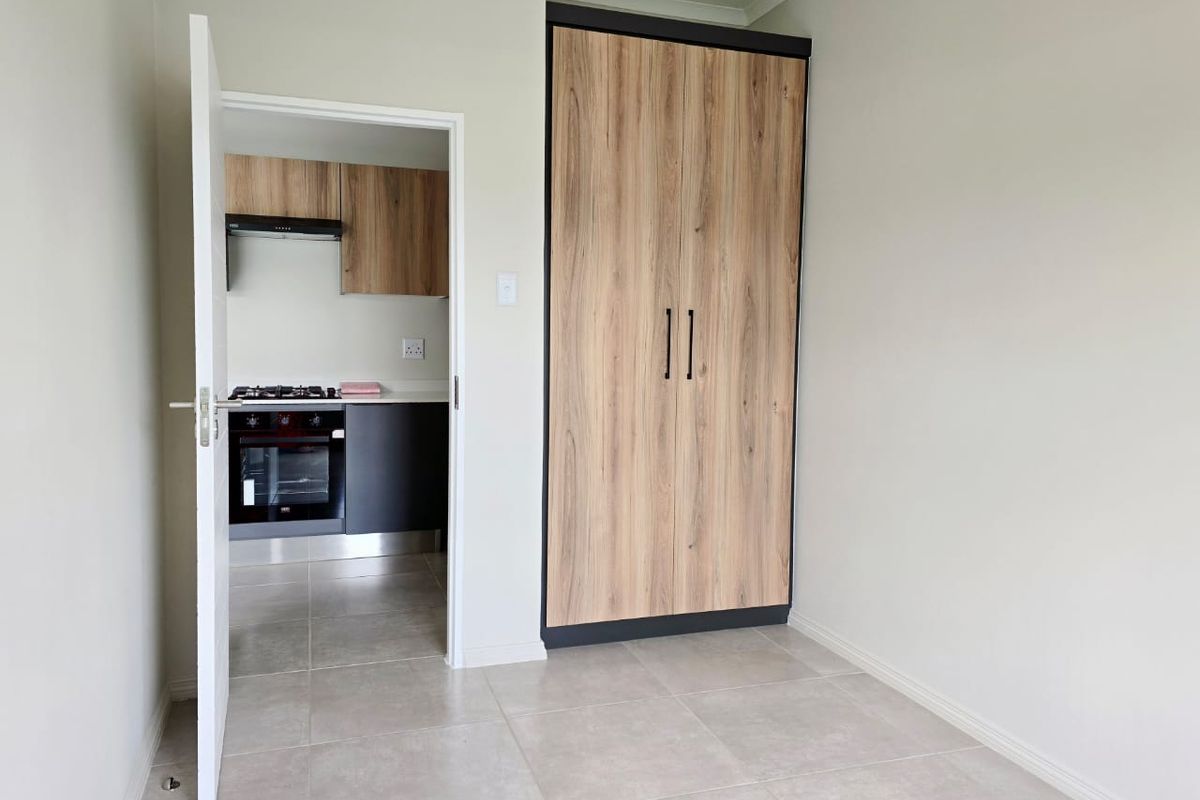 A bright, empty bedroom featuring modern wood-look built-in wardrobes, neutral tiled flooring, and a doorway leading to the kitchen area.