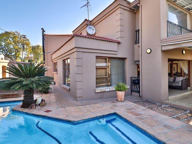 A well-maintained outdoor pool area featuring a paved deck, swimming pool, and access to a covered patio and upper-level balcony.