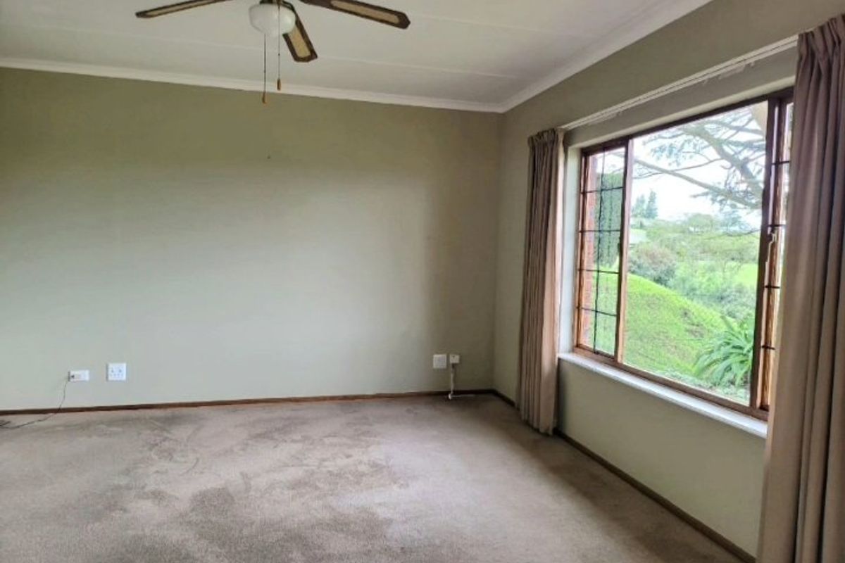 A spacious, empty living room featuring neutral walls, a ceiling fan, and a large window with views of the garden.