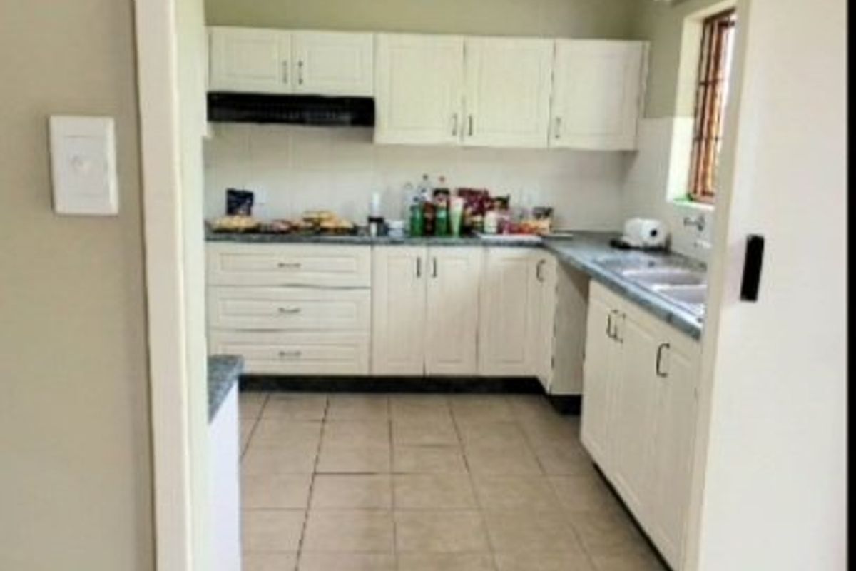 A functional kitchen featuring white built-in cabinetry, laminate countertops, a double sink, and tiled flooring, viewed from the adjacent doorway.