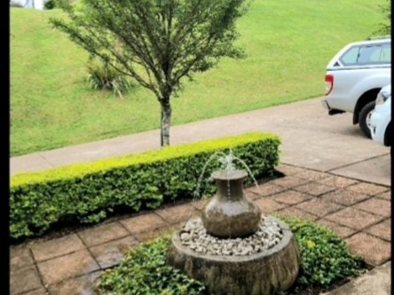 A landscaped garden area featuring a stone water fountain, paved pathway, and manicured hedge set against a grassy slope.