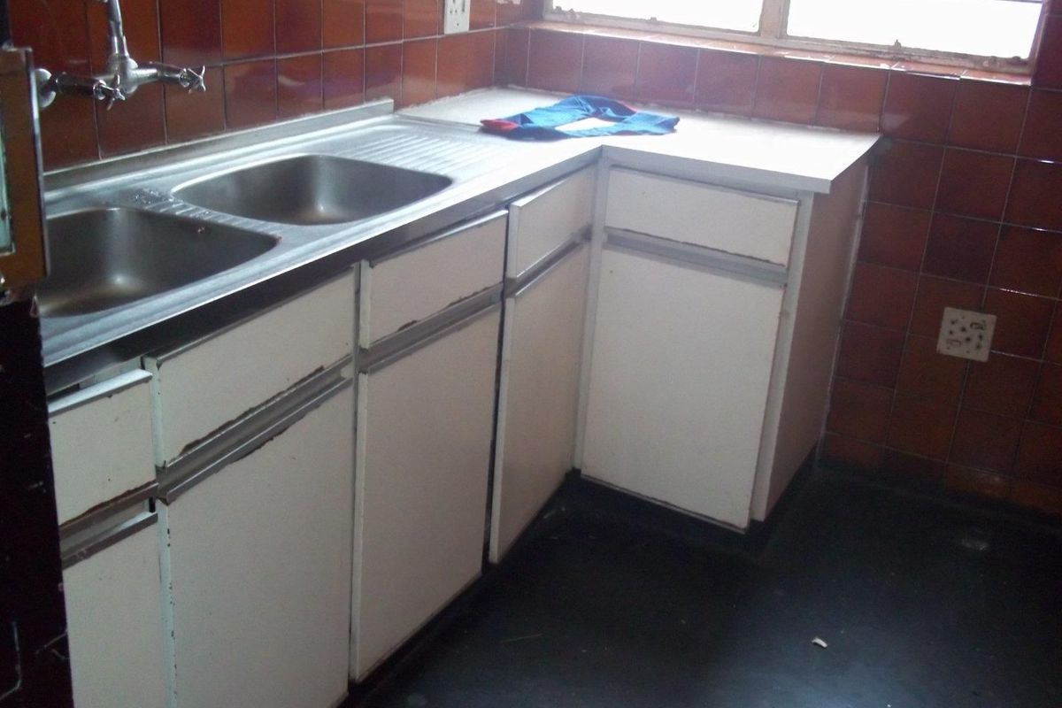 A dated kitchen area featuring white laminate cabinetry, a double bowl stainless steel sink, and brown ceramic wall tiles.