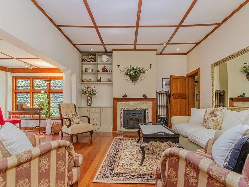 A cozy living room featuring a fireplace, built-in shelving, hardwood flooring, and distinctive timber ceiling beams with leaded glass windows.