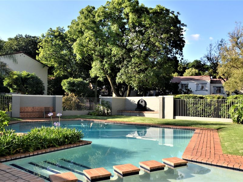 A well-maintained outdoor pool area featuring brick paving, a built-in water feature, and established garden surroundings within a secure townhouse complex.