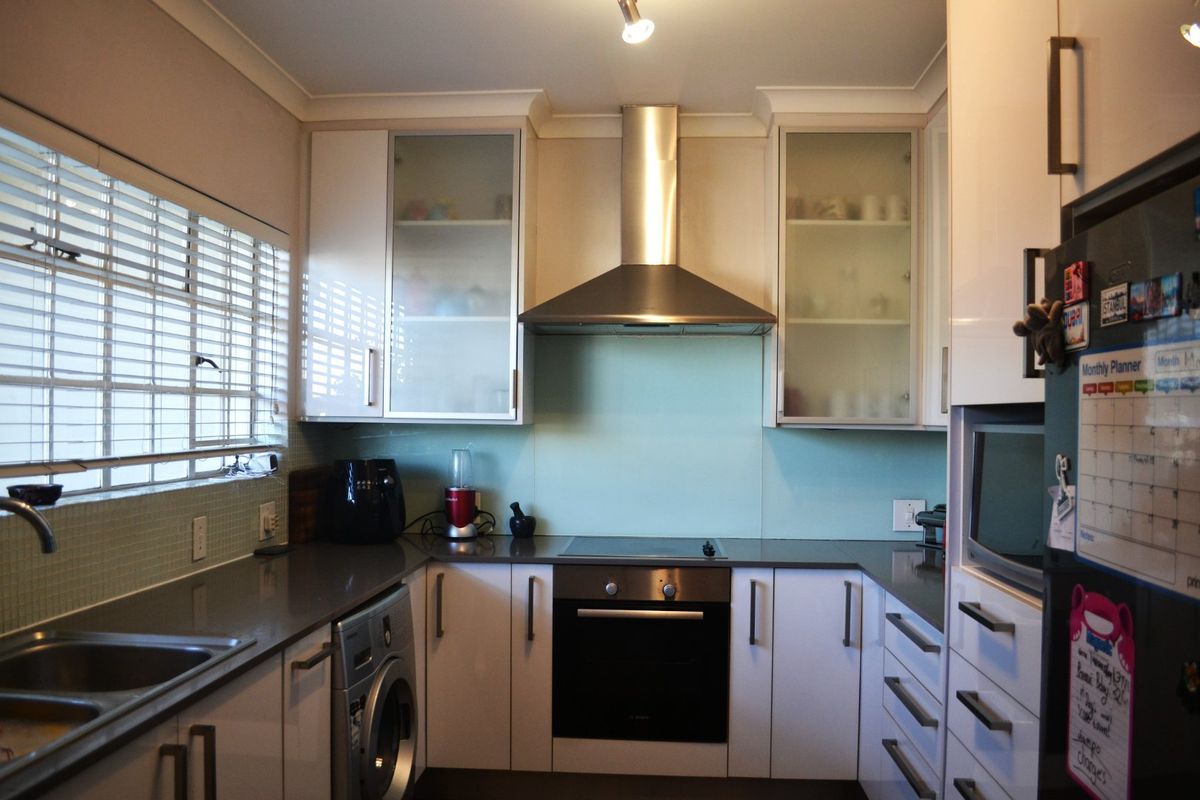 Modern kitchen featuring white cabinetry, stone countertops, glass splashback, stainless steel extractor fan, built-in oven, and an integrated washing machine.