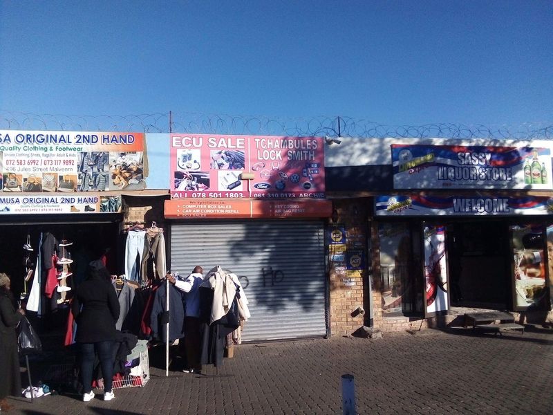 Commercial storefronts featuring security roller shutters, razor wire perimeter fencing, and a paved forecourt area in a busy street-level retail setting.