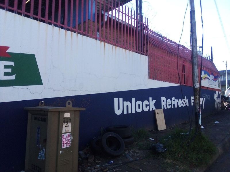 Exterior view of a commercial property boundary wall featuring security fencing and a utility transformer box, showing signs of significant wear and weathering.