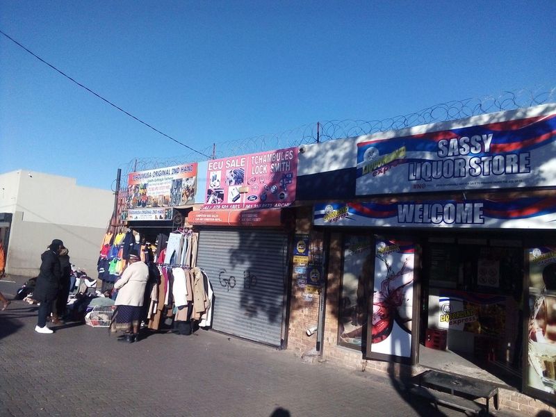 Street-level view of a commercial retail strip featuring multiple storefronts with security roller shutters and prominent signage.