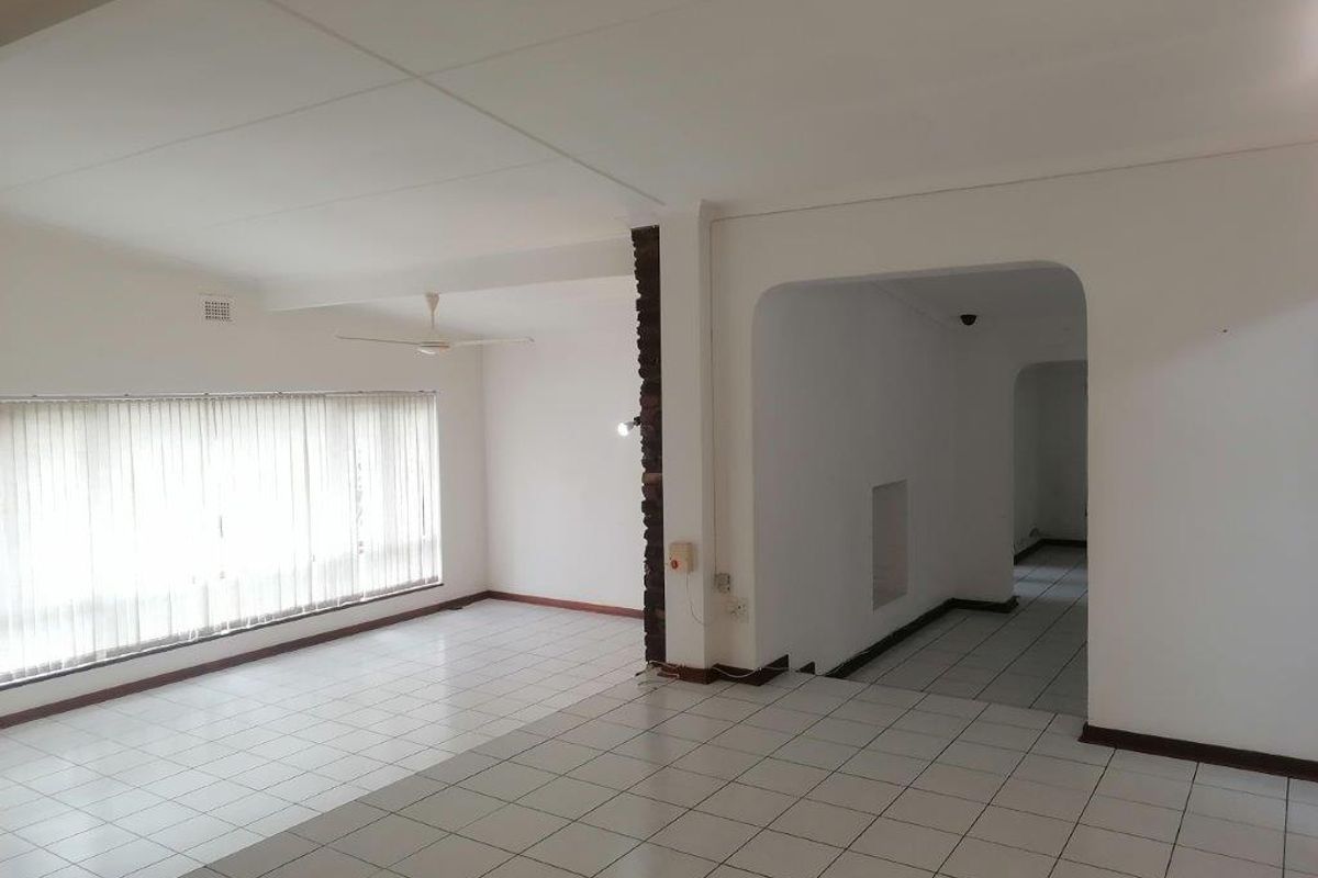 Spacious, empty living area featuring tiled flooring, an exposed brick pillar, and an archway leading to a hallway.