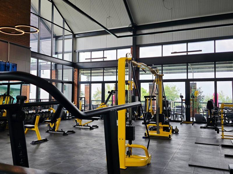 A modern, well-lit gym facility featuring high vaulted ceilings, exposed brick accents, and expansive glass walls providing ample natural light and outdoor views.