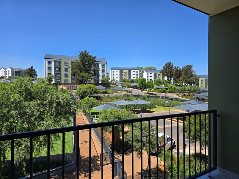 Elevated view from a balcony overlooking a well-maintained residential apartment complex with landscaped grounds, paved pathways, and shaded parking areas.