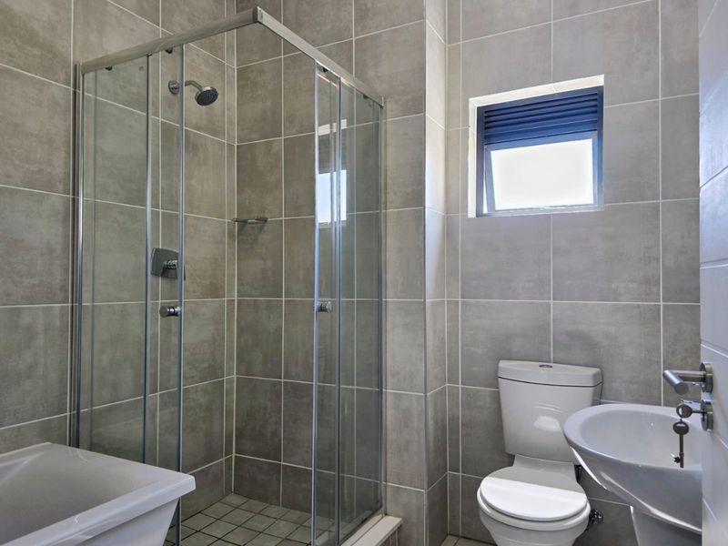 Modern bathroom featuring a glass-enclosed shower, pedestal basin, and toilet, finished with neutral grey wall and floor tiles.