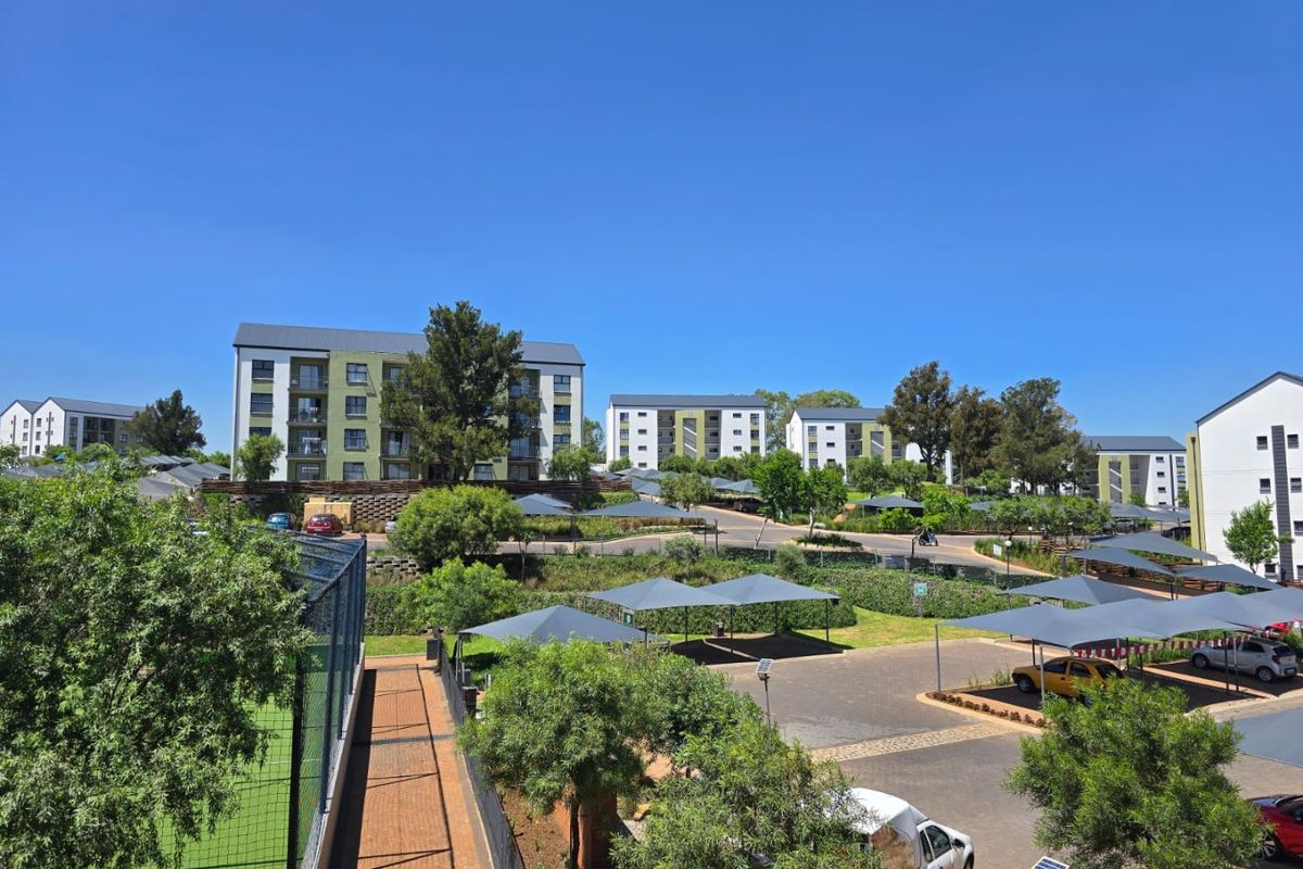 An elevated aerial view of the apartment complex grounds, featuring paved parking with shade cloth carports, landscaped areas, and a fenced sports court.