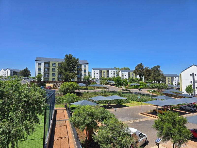 An elevated aerial view of the apartment complex grounds, featuring paved parking with shade cloth carports, landscaped areas, and a fenced sports court.