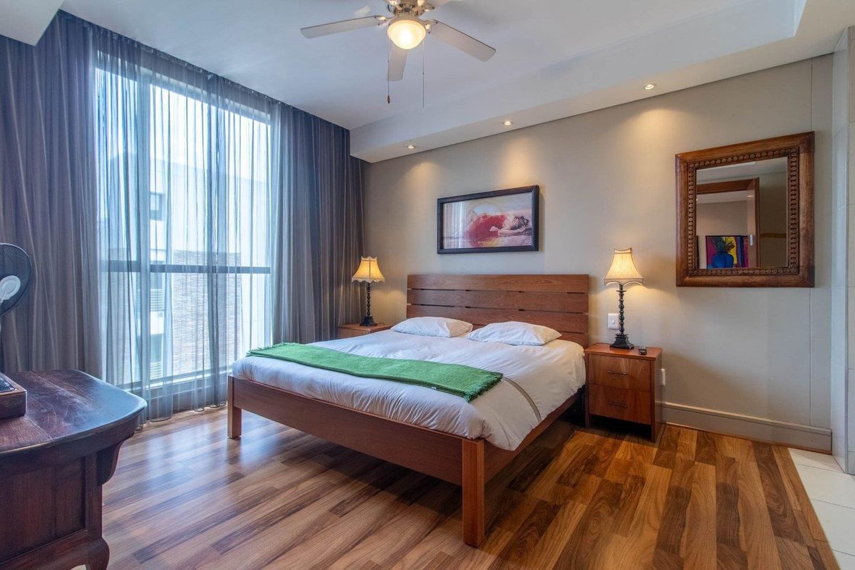 A well-maintained bedroom featuring warm-toned laminate flooring, a ceiling fan, recessed lighting, and a large floor-to-ceiling window with sheer curtains.