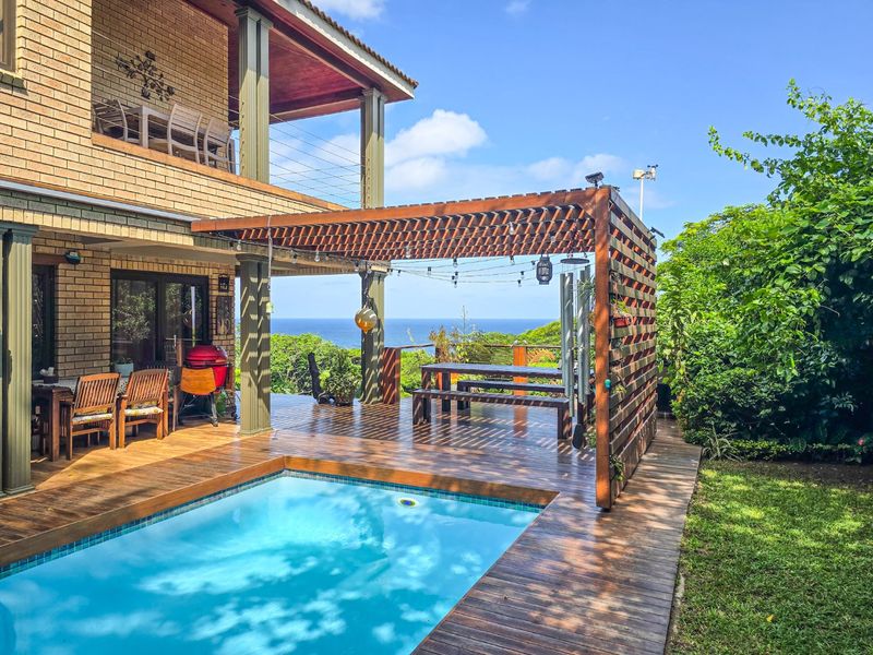 Outdoor pool area featuring timber decking, a wooden pergola, and a brick house facade with a balcony overlooking the ocean.